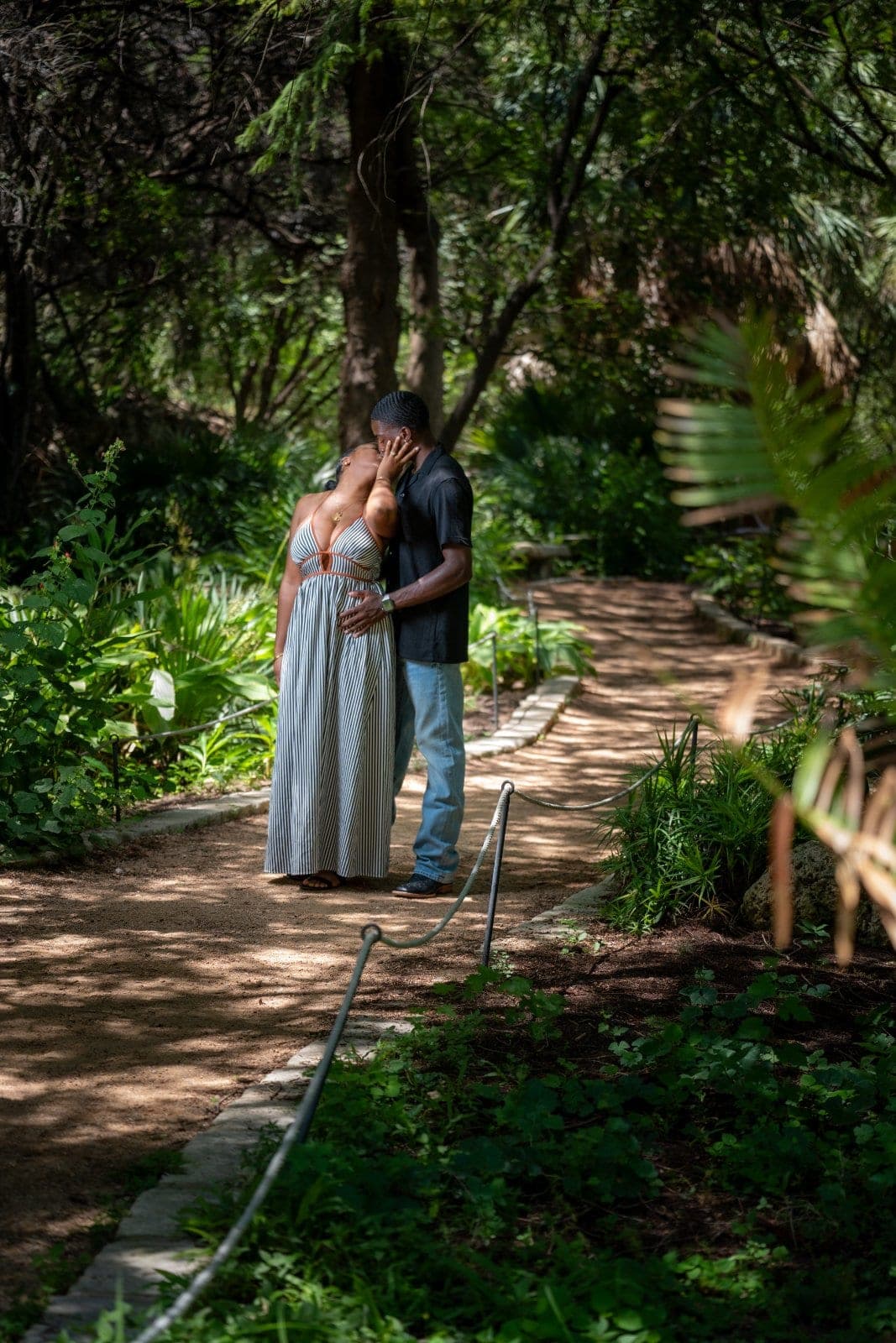 Newly engaged couple sharing a kiss on a tree-lined garden path during their Austin engagement photography session.